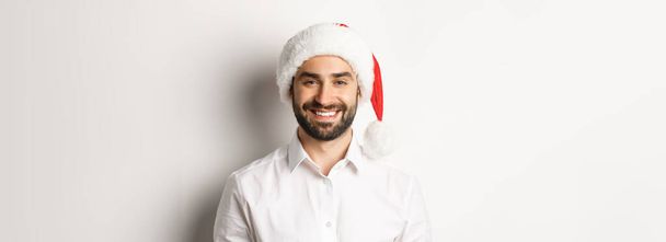 Close-up de homem barbudo feliz comemorando o Natal, vestindo chapéu de festa de Papai Noel e sorrindo, de pé sobre fundo branco. - Foto, Imagem