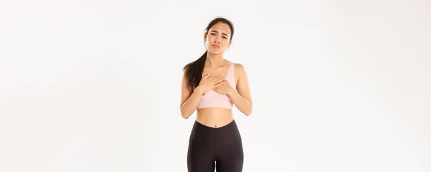 Sport, wellbeing and active lifestyle concept. Gloomy complaining asian fitness girl longing for something, holding hands on heart and sulking jealous, feel regret or upset, white background. - Photo, image