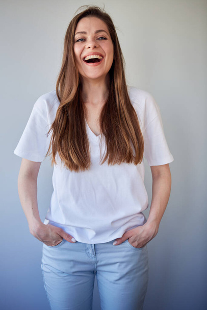 Laughing woman in white shirt keeps hands in pockets. Happy girl with long brown hair, isolated portrait. - Foto, imagen