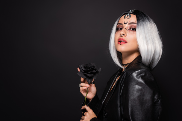 woman in witch halloween makeup and leather jacket posing with dark rose isolated on black - Photo, Image