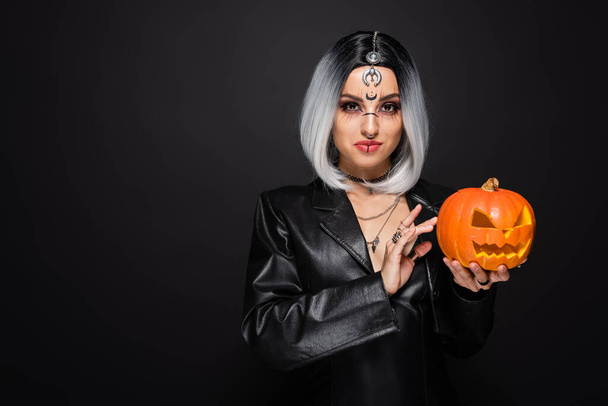 smiling woman in leather jacket and witch makeup holding carved pumpkin isolated on black - Photo, Image