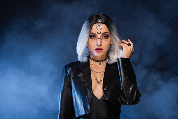 sexy ash blonde woman in leather jacket and halloween makeup looking at camera on dark background with blue fog - Фото, изображение