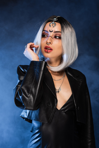 sexy ceniza rubia mujer en bruja maquillaje mirando hacia otro lado en fondo oscuro con humo azul - Foto, Imagen