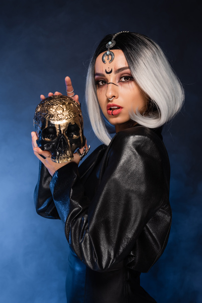 sexy witch style woman with creepy golden skull looking at camera near blue fog on dark background - Photo, Image