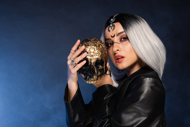 woman in witch makeup holding golden skull on dark background with blue fog - Fotó, kép