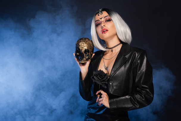 woman in witch halloween costume holding golden skull and black rose on dark background with blue smoke - Fotó, kép