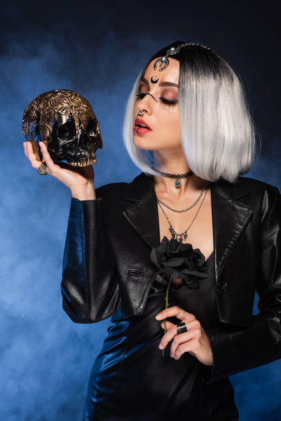 sexy woman in witch costume holding black rose and looking at golden skull on dark background with blue fog - Zdjęcie, obraz