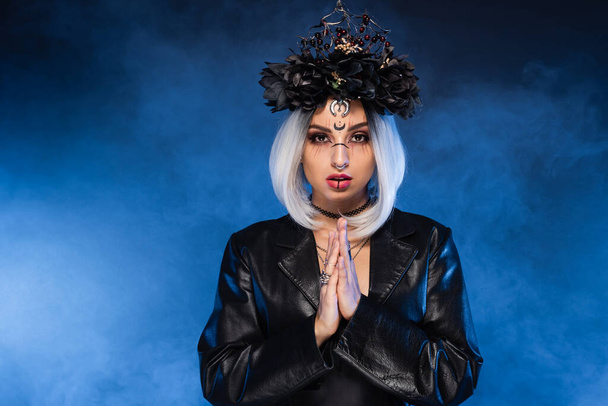 čarodějnice styl žena v černé věnec a kožené sako stojící s modlící se ruce na tmavém pozadí s modrou mlhou - Fotografie, Obrázek