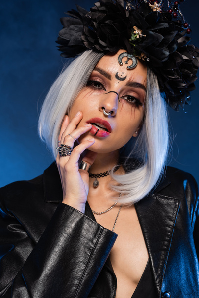 sexy woman in witch makeup and black wreath with accessories looking at camera on blue background - Zdjęcie, obraz