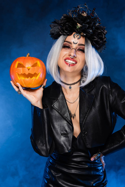Cadı kostümü giymiş neşeli bir kadın ve elinde oyulmuş balkabağı, mavi sisin yanında. - Fotoğraf, Görsel