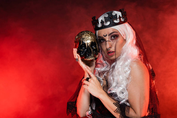 mulher loira estilo bruxa em coroa preta e véu olhando para a câmera perto de crânio dourado no fundo fumegante vermelho - Foto, Imagem