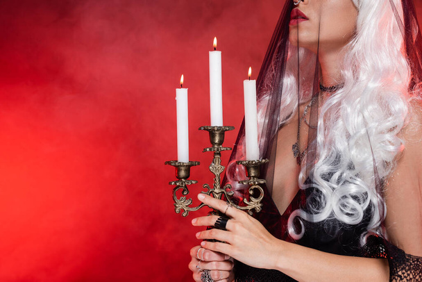cropped view of blonde witch in black veil holding burning candles on background with red smoke - 写真・画像