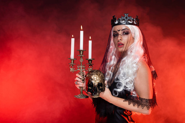 blonde woman in witch costume holding golden skull and burning candles on dark background with smoke - Photo, Image
