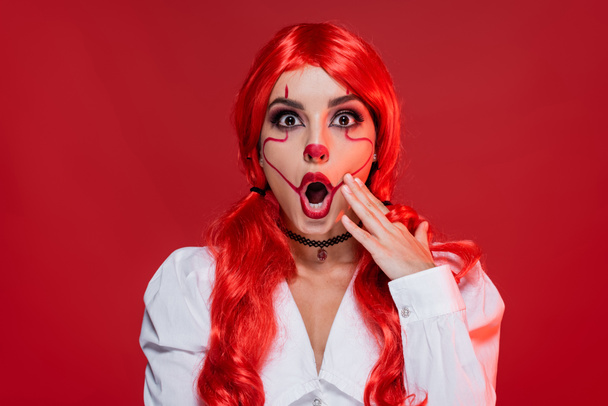 femme étonnée avec halloween maquillage toucher le visage et en regardant la caméra isolée sur rouge - Photo, image