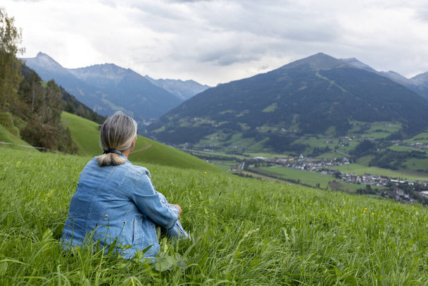 A gray-haired woman in a denim suit sits on a grassy slope and looks towards the mountains. Austria, Salzburg. High quality photo - Photo, Image