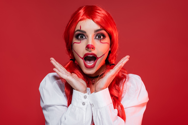 amazed redhead woman with halloween makeup holding hands near face isolated on red - Foto, Bild