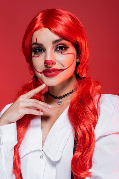 портрет улыбающейся женщины с клоунским макияжем и яркими волосами, касающимися подбородка и смотрящей на камеру, изолированную на красном - Фото, изображение