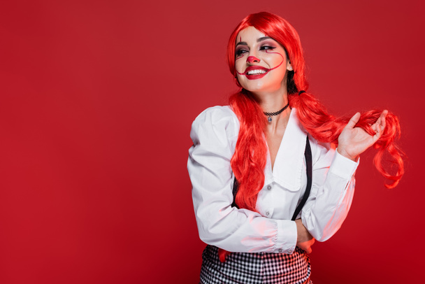 joyful redhead woman in clown makeup and white blouse looking away isolated on red - Фото, изображение