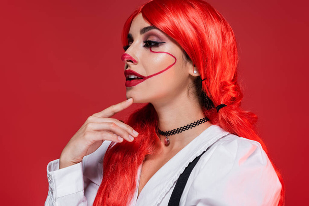 přemýšlivá a usmívající se žena s jasnými vlasy a klaun make-up odvrací izolované na červené - Fotografie, Obrázek