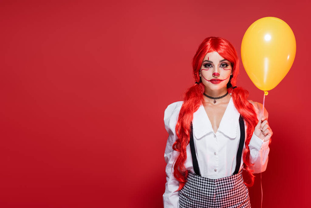 jeune rousse femme en chemisier blanc et maquillage de clown tenant ballon jaune isolé sur rouge - Photo, image