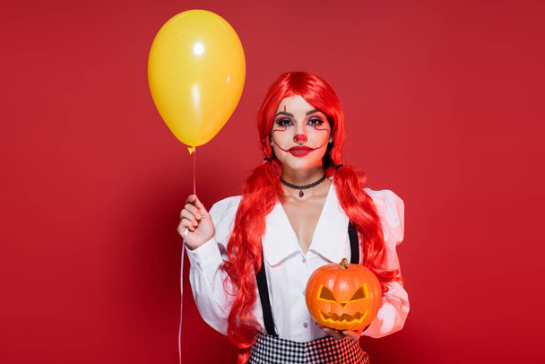 woman with clown makeup and bright hair holding yellow balloon and carved pumpkin isolated on red - Foto, afbeelding