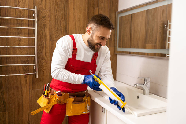 Professionele loodgieter, mannelijke werknemer in uniform installeren wastafel en waterleiding in nieuw appartement. - Foto, afbeelding