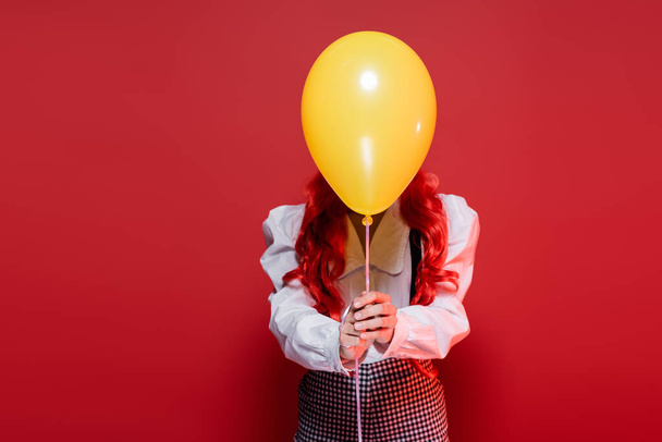mujer joven con el pelo de color que oscurece la cara con globo amarillo en la fiesta de halloween aislado en rojo - Foto, imagen