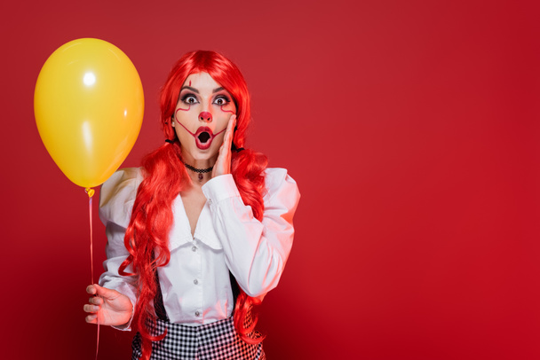 amazed redhead woman in clown makeup holding yellow balloon and touching face isolated on red - Fotó, kép