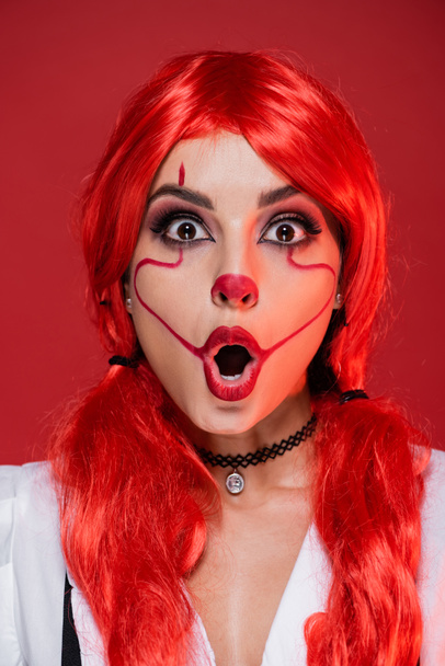pelirroja mujer asombrada con el maquillaje de Halloween mirando a la cámara aislada en rojo - Foto, Imagen