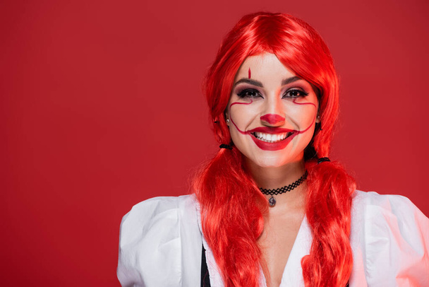 radosna kobieta z jasnymi kolorowymi włosami i makijażem klauna patrząc na kamerę odizolowaną na czerwono - Zdjęcie, obraz
