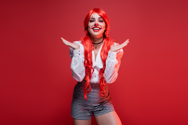 šťastná žena s dlouhými barevnými vlasy a klaun make-up ukazuje s rukama izolovaných na červené - Fotografie, Obrázek