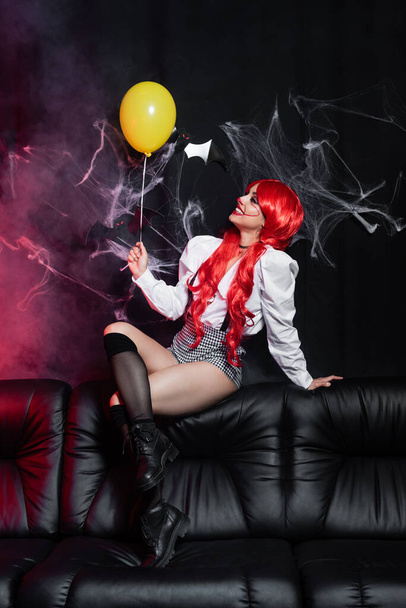 sexy woman with red hair and clown makeup sitting with yellow balloon on leather couch near spiderweb on black background - Foto, imagen