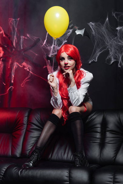 sexy woman in clown makeup and black knee socks holding yellow balloon near spiderweb on dark background - Photo, image