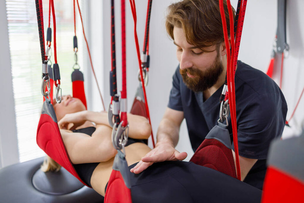 Physiotherapist assists female patient undergoing rehabilitative physiotherapy on a suspension rope system. Modern clinic using advanced rehabilitating therapy - Foto, imagen