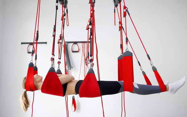 Young female patient hanging on modern suspensions system at rehabilitation center. Therapeutic exercises and neuromuscular activation on red cord slings - Фото, изображение