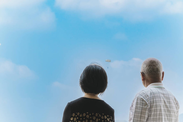 Back view of Asian parents looking with worry and concern during parachute training from airplane for army cadet with blurred image of parachute and sky in background. Family relationship concept. - Fotografie, Obrázek