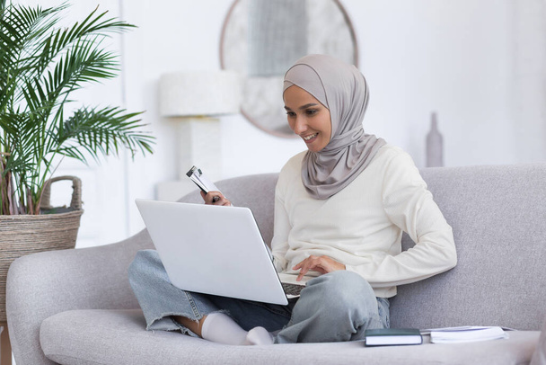 Young beautiful muslim woman in gray hijab studying at home sitting on sofa, female student using laptop for distance learning, watching lecture in living room. - Foto, immagini