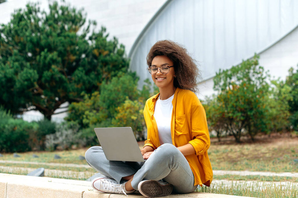 Modern lovely positive clever african american curly haired young woman, female student, freelancer, in stylish casual clothes, sitting with a laptop outdoors, looking at the camera, smiling happily - Valokuva, kuva