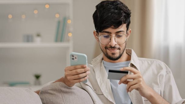 Lähikuva parrakas arabialainen liikemies lasit luottokortilla ja puhelin tekee ostoksia verkossa ostoksia tulee pankkitunnus verkkokaupan käyttää mobiili taloudellinen sovellus varaus järjestyksessä ostaminen - Valokuva, kuva
