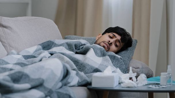 Indischer bärtiger trauriger, erschöpfter Mann, der auf der heimischen Couch liegt und krank mit einer Decke bedeckt ist und an Fieberkrankheiten leidet, Coronavirus-Konzept betrachtet Medikamente auf dem Tisch, die der Behandlung von Covid19 saisonaler Grippe müde sind - Foto, Bild