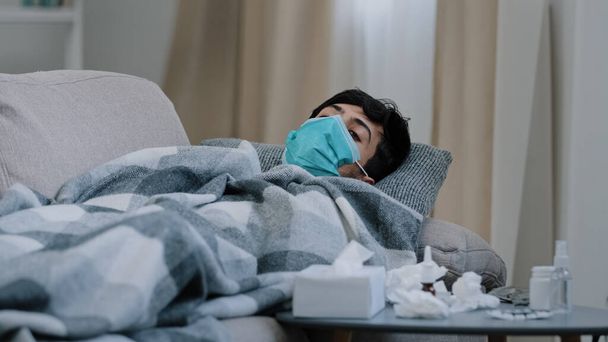 Sick guy man suffering from illness feeling unwell flu coronavirus symptoms wears medical mask lies at home sofa covered with blanket looking at table with medications waves head no hiding under plaid - Photo, Image