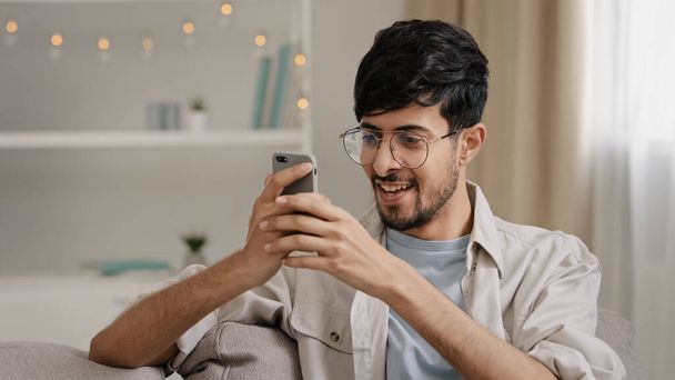 Junger Mann in den 30er Jahren, bärtiger Mann mit Brille sitzt zu Hause auf der Couch und schaut auf das Handy und macht eine Geste, die ihm eine gute Nachricht bringt. - Foto, Bild