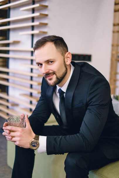 A young man with a beard, dressed in a classic business suit, sitting in the hands of a glass of whiskey. Portrait of a man relaxing with a glass of hard liquor. - Фото, изображение