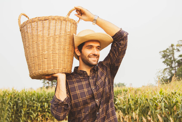 Alegre campesino miró al maizal con sonrisa y cargó canasta en su hombro trabajando duro y caminando orgullosamente para recoger los cultivos que plantó en el campo de maíz. - Foto, Imagen