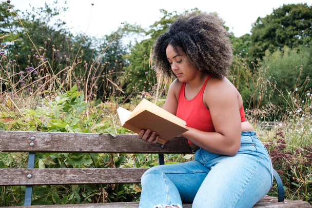 Young mixed race woman is reading a book seated in a bench in a park. Curly hair and she is wearing red t-shirt and jeans. Chubby body build. - 写真・画像