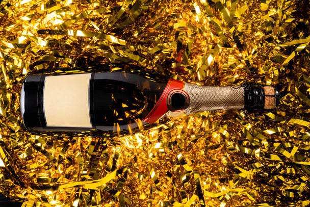 Top view of a corked bottle of champagne on shiny golden tinsel ribbons. Gold sparkle serpentine, stripes of glitter sequins. Festive background for the new year, holiday and birthday. Screensaver for - Photo, Image