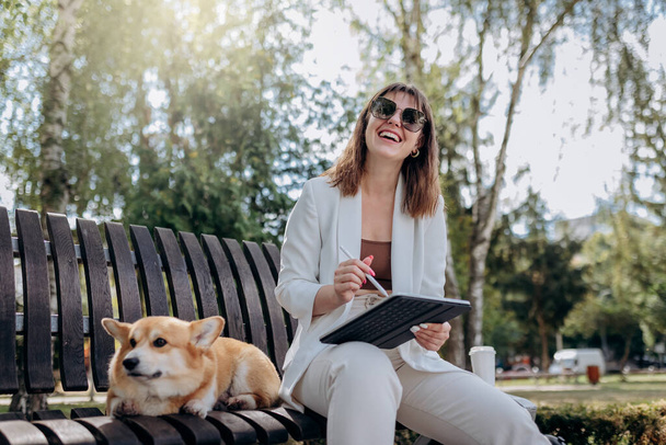 Pretty businesswoman in white suit sitting in city parkland with Welsh Corgi Pembroke dog and working on digital tablet - 写真・画像