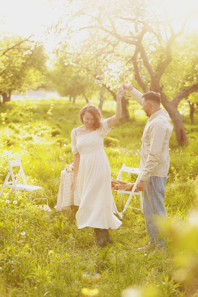 Young couple enjoying food and drinks in beautiful summer green park on romantic date picnic, handsome man and woman with curly hair are hugging outdoors. - 写真・画像