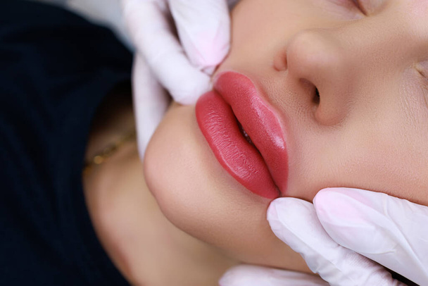 procedure of permanent make-up of the lips with the help of a machine, the master stretches the corners of the lips of the model - Photo, Image
