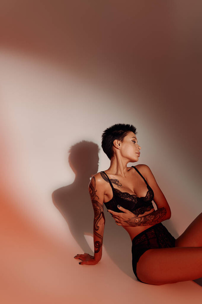 tetovált barna nő szexi fehérnemű ül a piros háttér árnyék - Fotó, kép